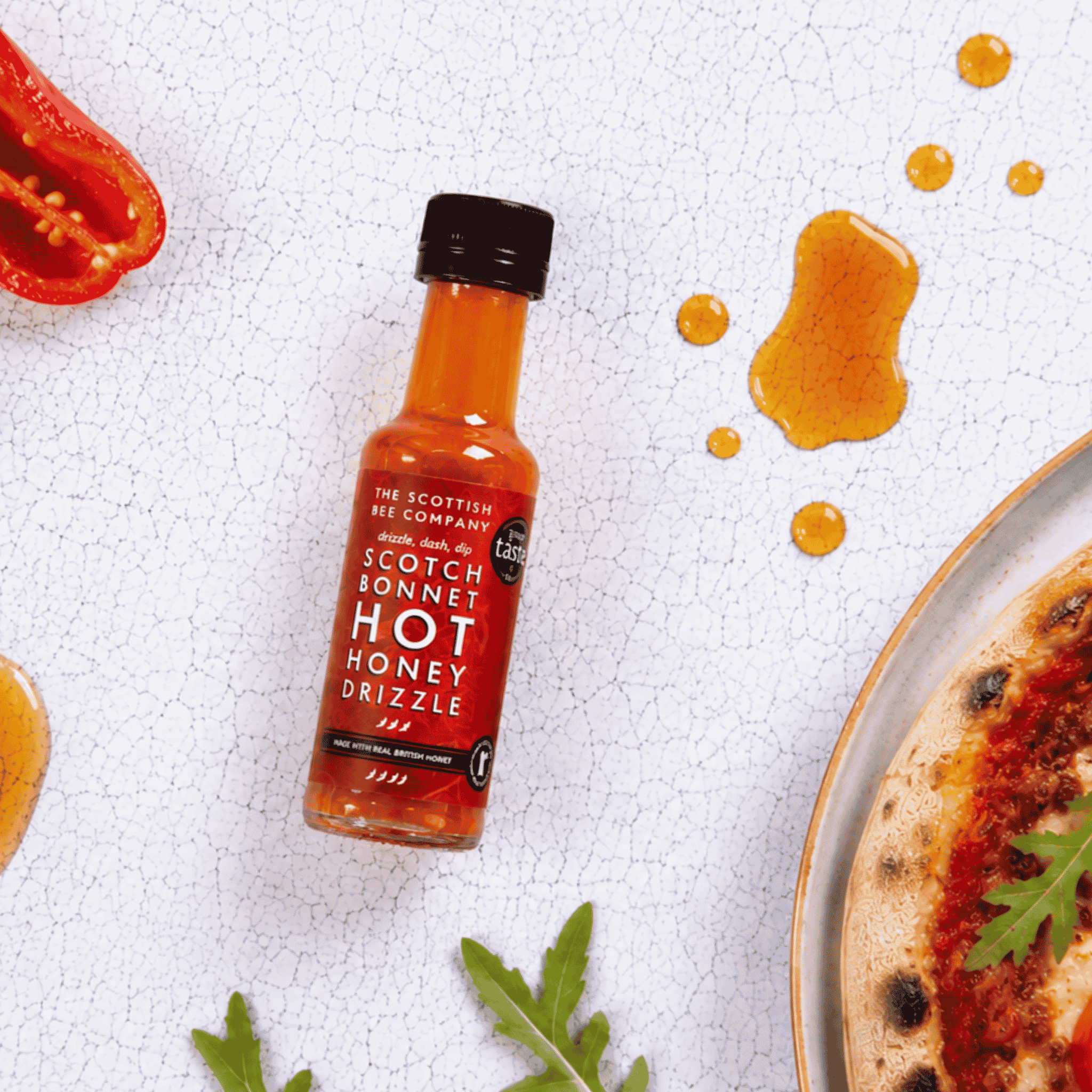 A bottle of The Scottish Bee Company Scotch Bonnet Hot Honey Drizzle sauce, featuring a red label and black cap, styled on a white textured surface with honey drizzles, a red pepper slice, and a pizza edge.