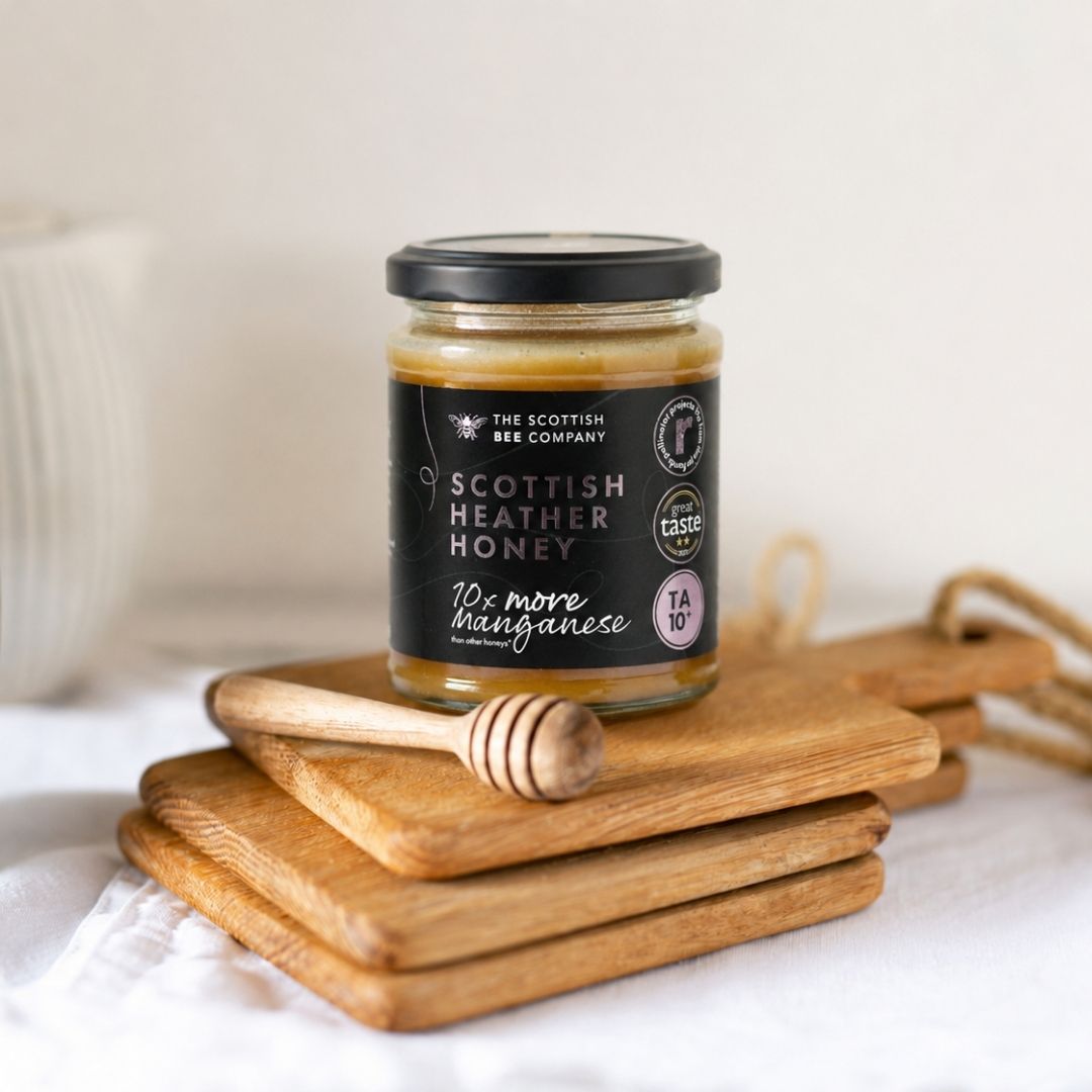 A glass jar of Scottish Heather Honey from The Scottish Bee Company sits on a stack of wooden cutting boards with a honey dipper, against a soft, neutral background.