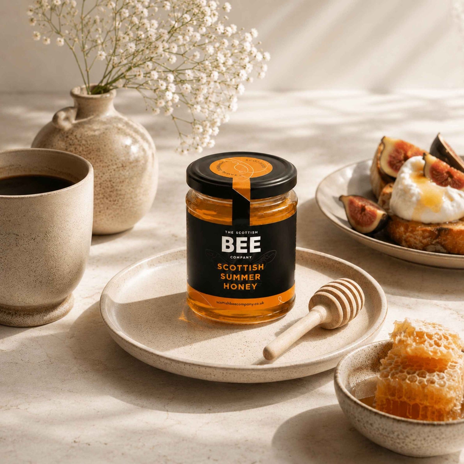A jar of Scottish Summer Honey from Bee Gardens sits on a ceramic plate, styled with a honey dipper, a cup of coffee, honeycomb, figs, and a vase of baby&