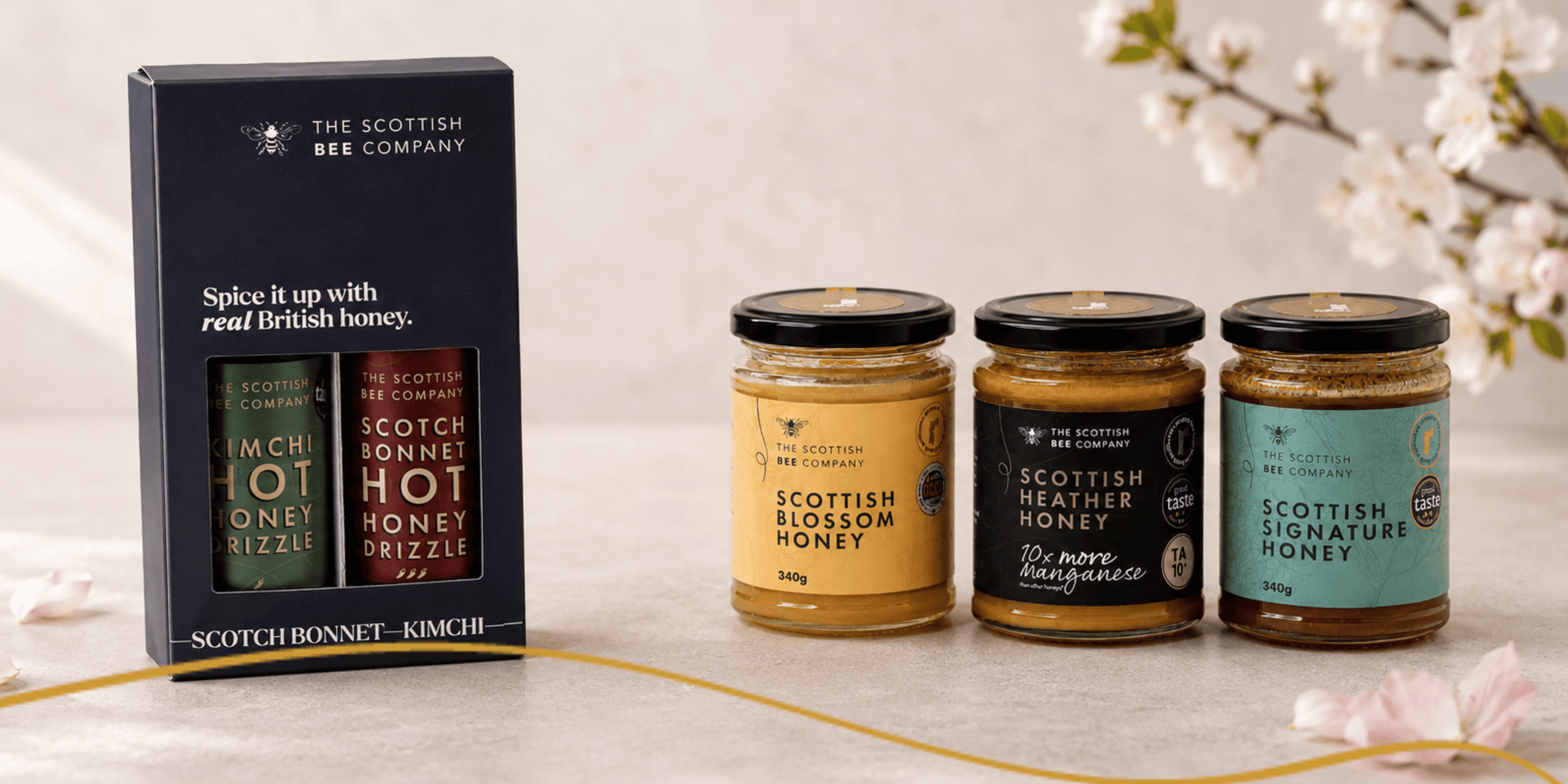 The Scottish Bee Company gift set and three jars of Scottish Blossom Honey, Scottish Heather Honey and Scottish Signature Honey styled on a neutral premium background with soft pink petals.