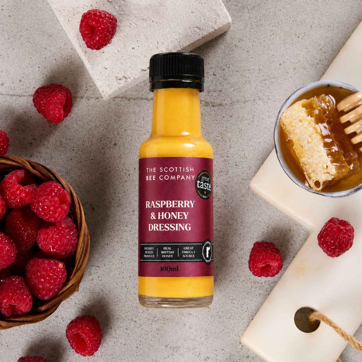 A bottle of The Scottish Bee Company Raspberry &amp; Honey Dressing is styled in a flat-lay arrangement with fresh raspberries, a bowl of honey, and stone tiles on a textured gray background.