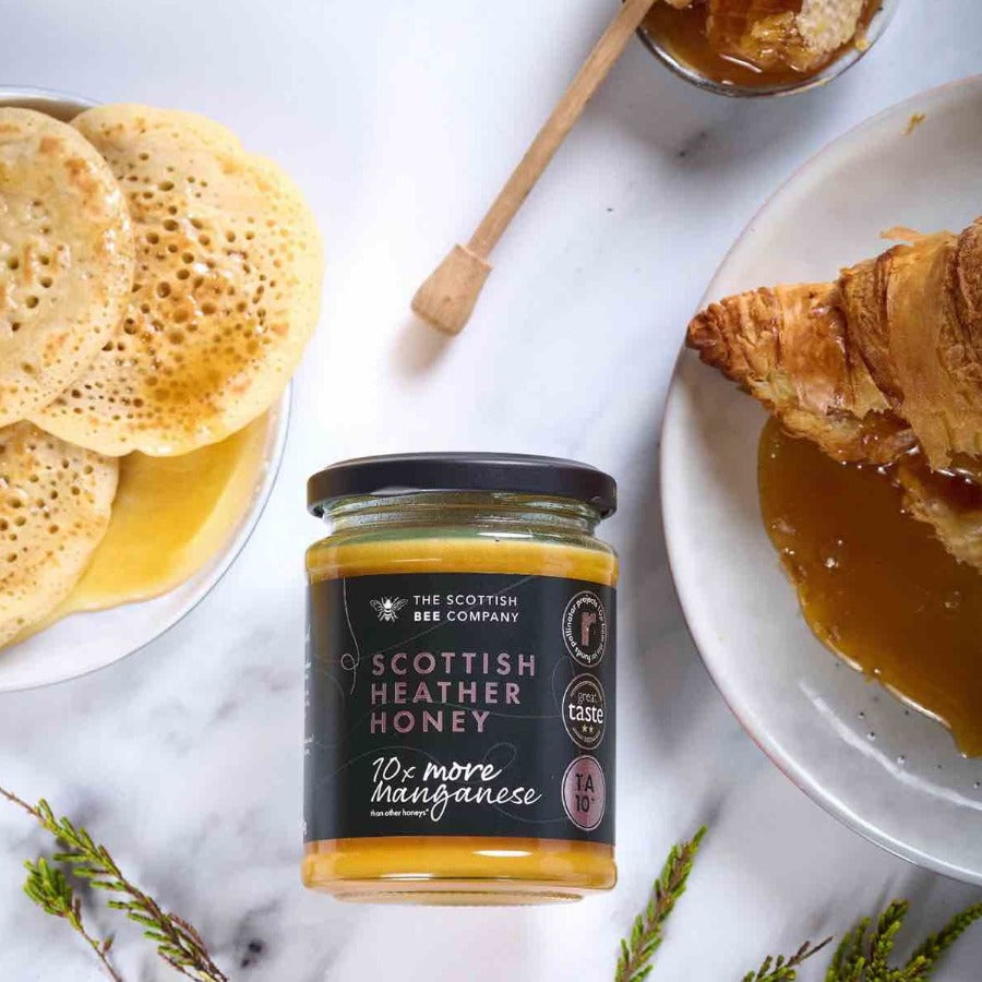 Jar of Scottish Heather Honey from The Scottish Bee Company with pancakes and croissants on a marble surface.