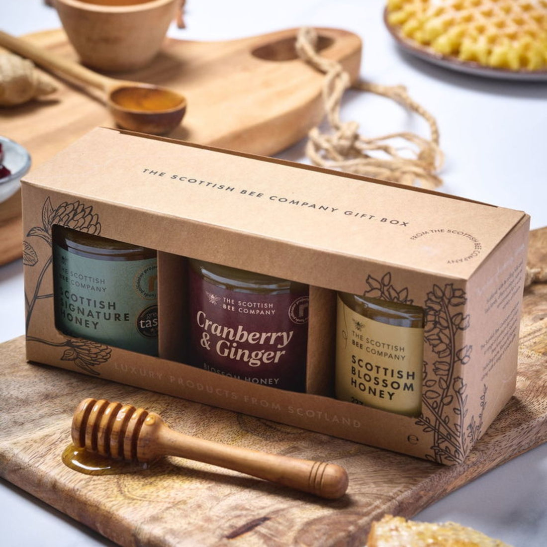 Scottish Bee Company honey gift set on a wooden surface with a honey dipper. Includes Scottish Signature Honey, Cranberry &amp; Ginger Blossom Honey and Scottish Blossom Honey