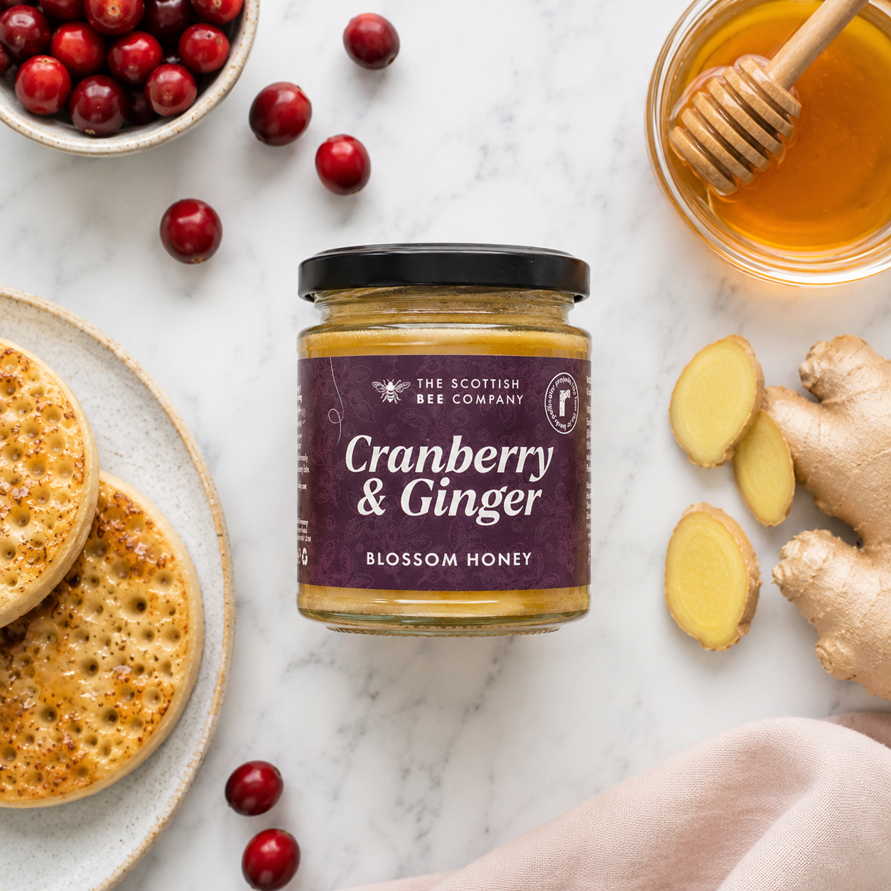 Jar of Cranberry &amp; Ginger Blossom Honey from The Scottish Bee Company on a marble surface with crumpets, honey, and ginger.