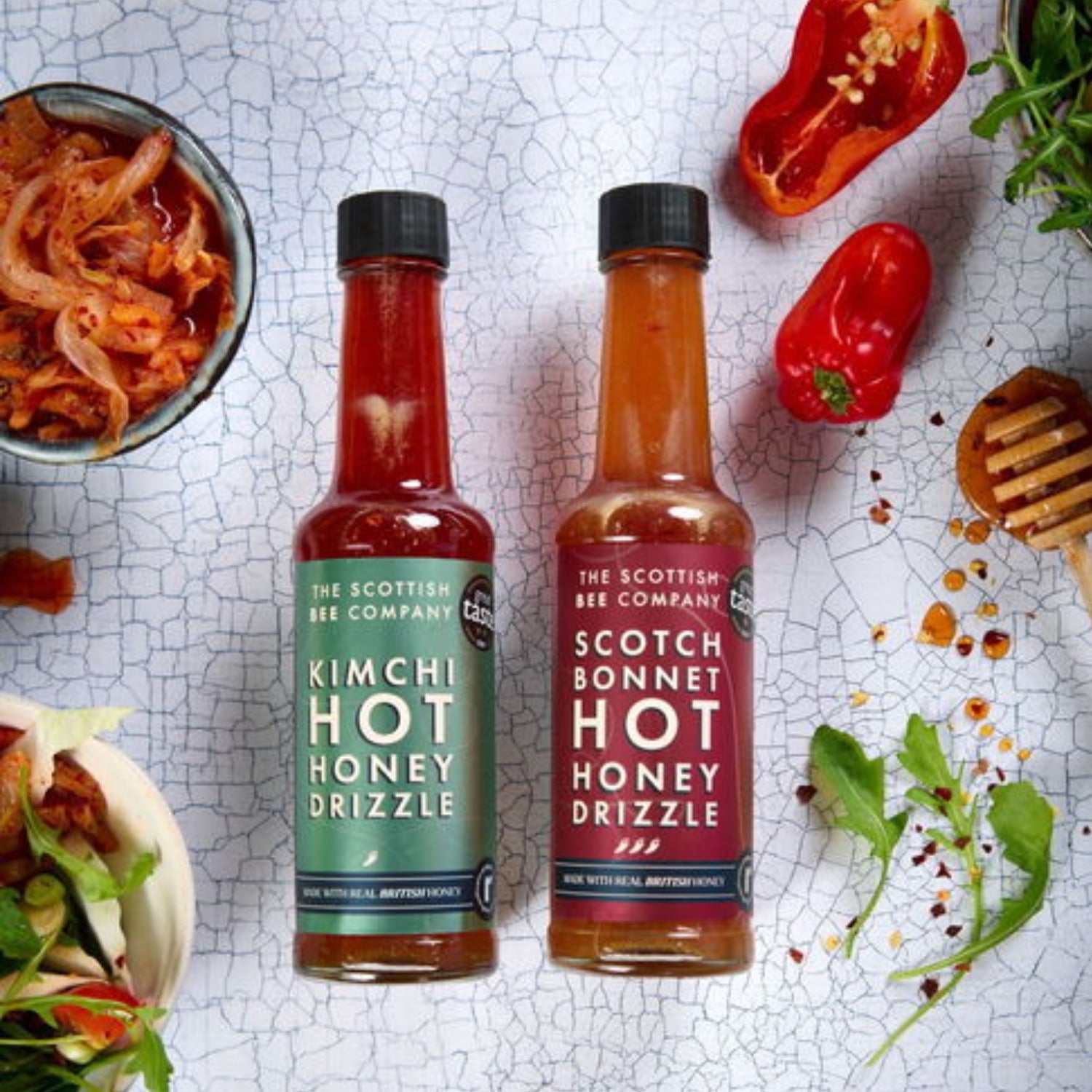 Two bottles of Scottish Bee Company hot honey drizzles on a textured surface with food ingredients. Contains Kimchi Hot Honey Drizzle and Scotch Bonnet Hot Honey Drizzle