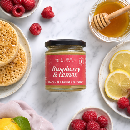 Jar of raspberry and lemon honey surrounded by raspberries, lemons, and crumpets on a marble surface.