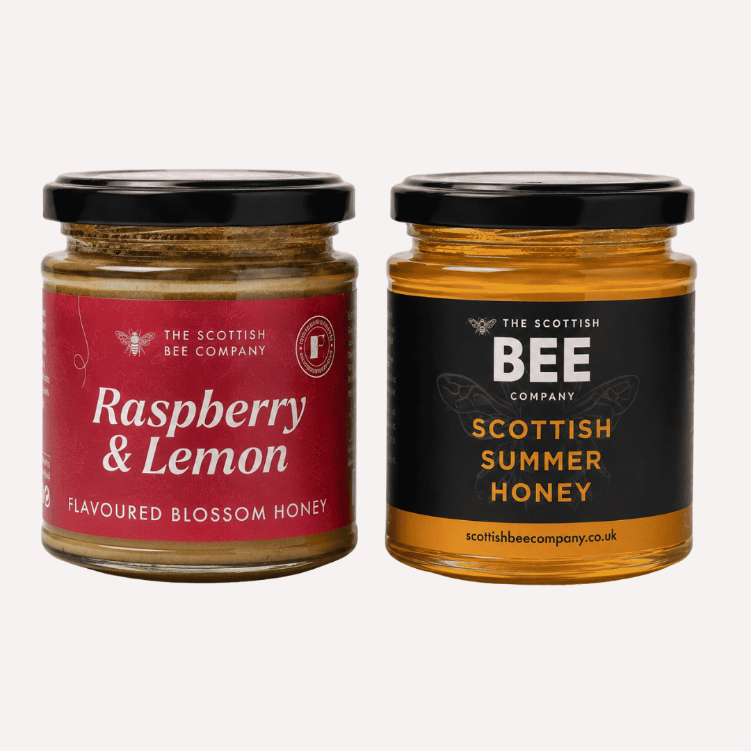Two glass jars of The Scottish Bee Company honey on a light gray background: Raspberry &amp; Lemon Flavoured Blossom Honey with a red label, and Scottish Summer Honey with a black label.