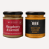 Two glass jars of The Scottish Bee Company honey on a light gray background: Raspberry & Lemon Flavoured Blossom Honey with a red label, and Scottish Summer Honey with a black label.