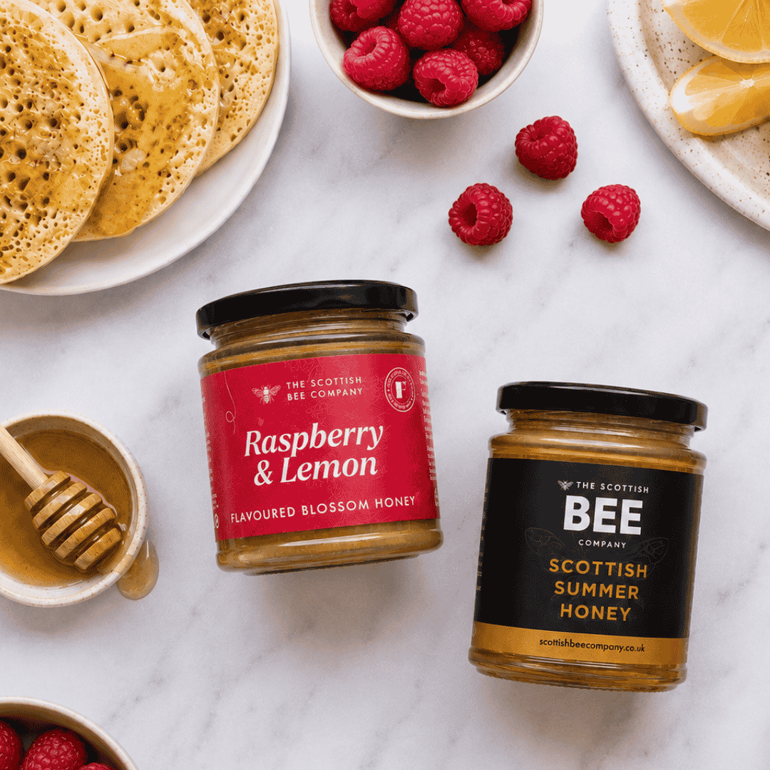 Two glass jars of honey from The Scottish Bee Company on a white marble surface, styled with raspberries, lemon slices, and pancakes. One jar has a red label for Raspberry &amp; Lemon Flavoured Blossom Honey, and the other has a black label for Scottish Summer Honey.