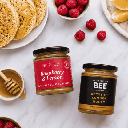 Two glass jars of honey from The Scottish Bee Company on a white marble surface, styled with raspberries, lemon slices, and pancakes. One jar has a red label for Raspberry &amp; Lemon Flavoured Blossom Honey, and the other has a black label for Scottish Summer Honey.