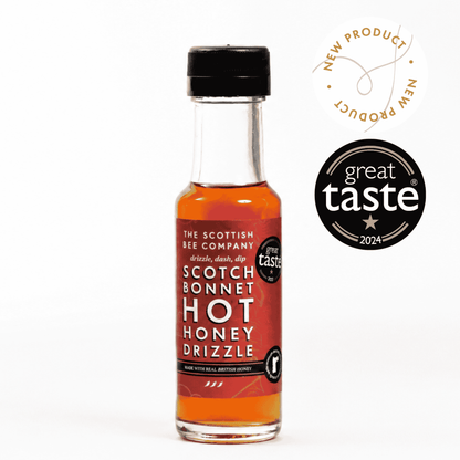 A small bottle of The Scottish Bee Company Scotch Bonnet Hot Honey Drizzle syrup with amber liquid and a red label, shown on a white background with a &
