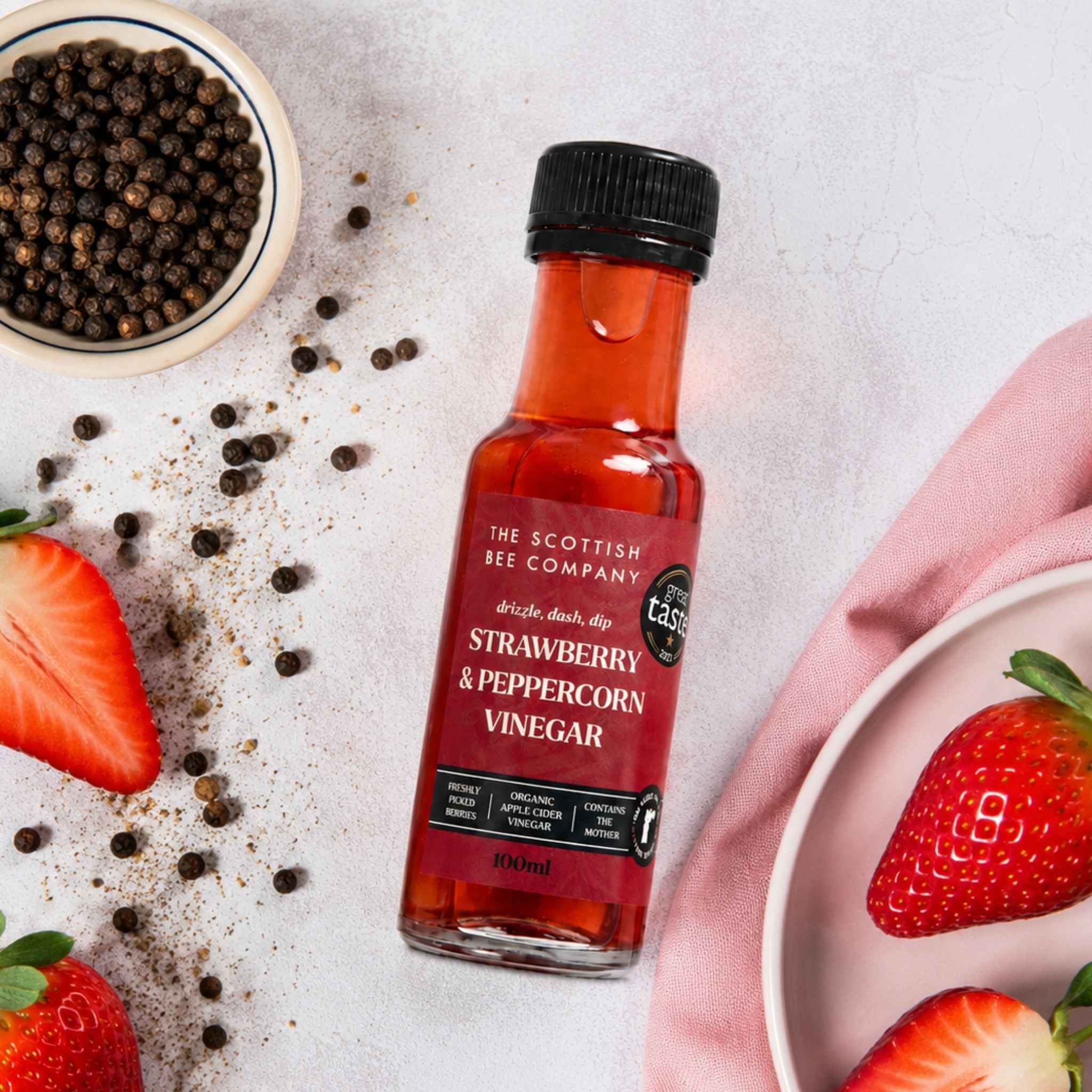 A bottle of The Scottish Bee Company Strawberry &amp; Peppercorn Vinegar is styled on a light textured surface with fresh strawberries, whole peppercorns, and a pink napkin.