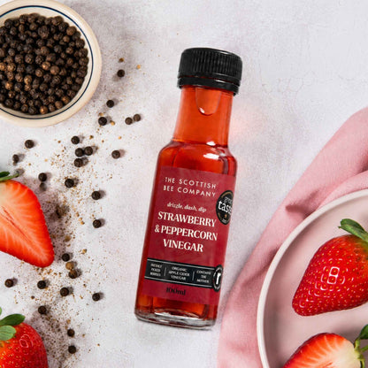 A bottle of The Scottish Bee Company Strawberry &amp; Peppercorn Vinegar is styled on a light textured surface with fresh strawberries, whole peppercorns, and a pink napkin.